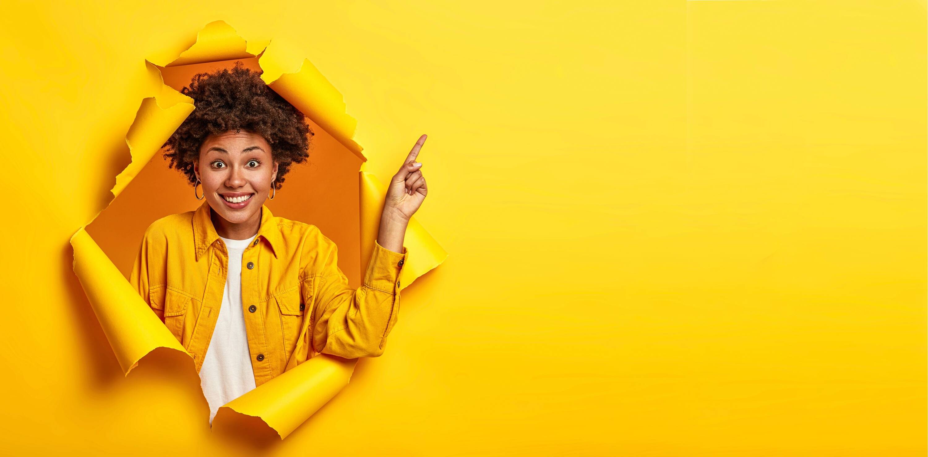 studio-shot-positive-african-american-woman-points-finger-copy-space-excited-by-good-information-smiles-pleasantly-wears-yellow-jacket-stands-ripped-paper-hole.jpg