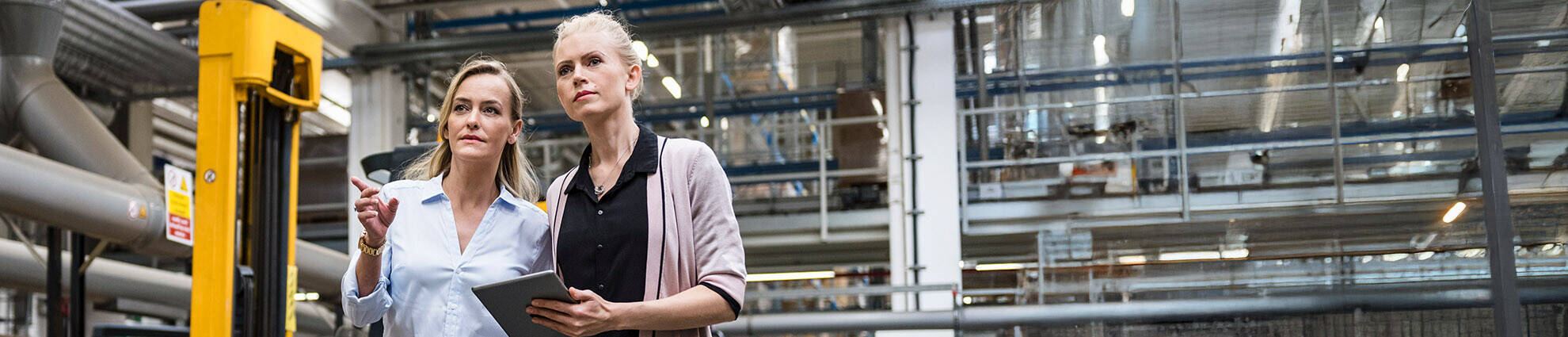 Two female professionals with a tablet are walking and talking on a factory shop floor.