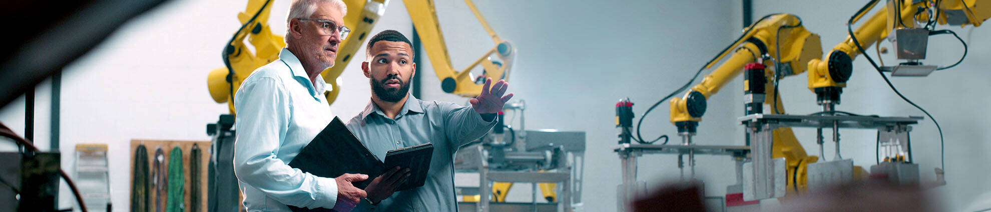 A young male professional pointing at machinery while discussing with a senior professional holding a tablet.