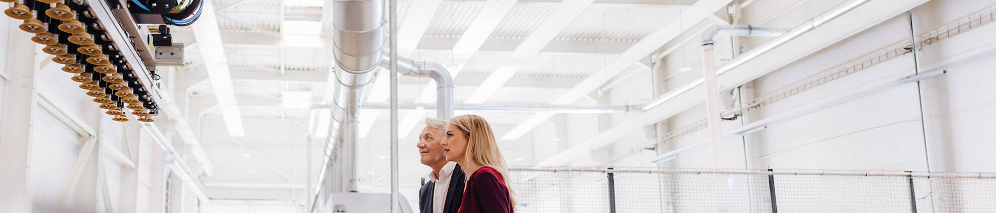 A senior male professional with a younger female professional looking at a machine in a factory.