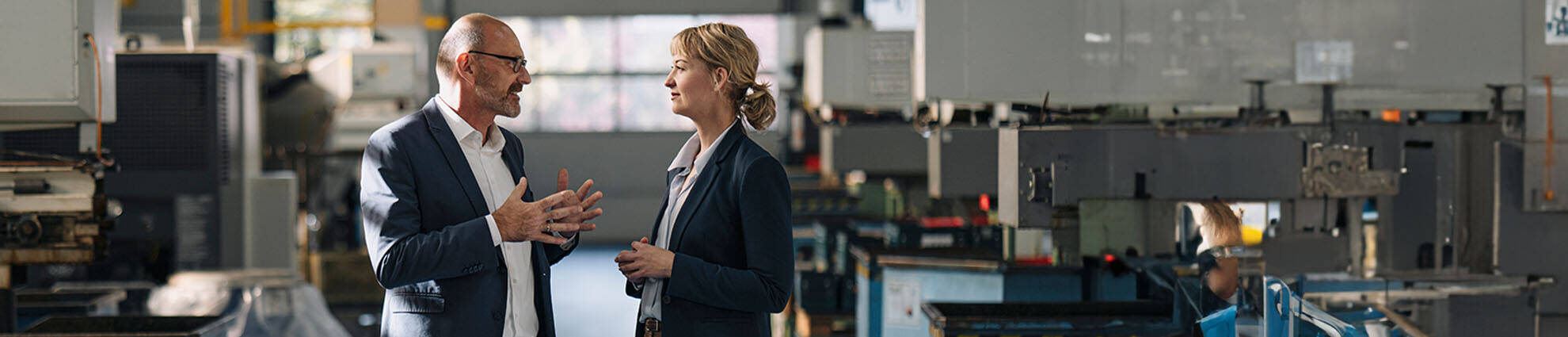 A male professional talking to a female professional, standing face to face, in a factory setting.