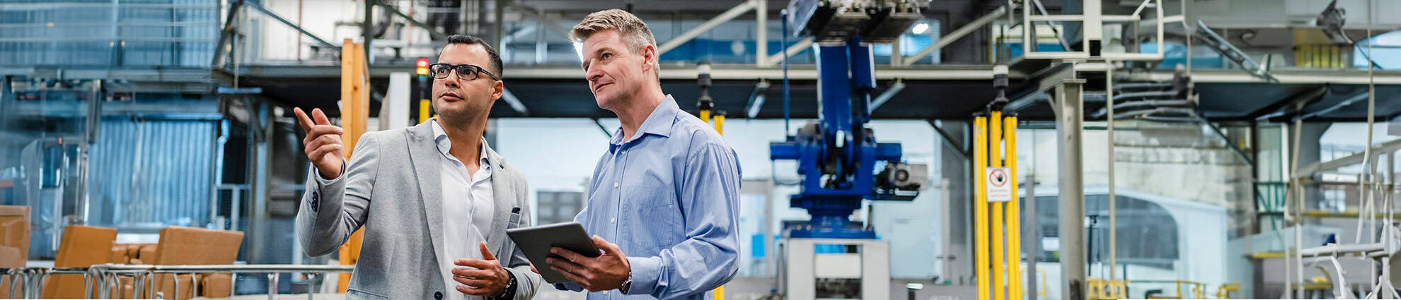 A male professional is pointing while talking with another male professional holding a tablet in a factory.