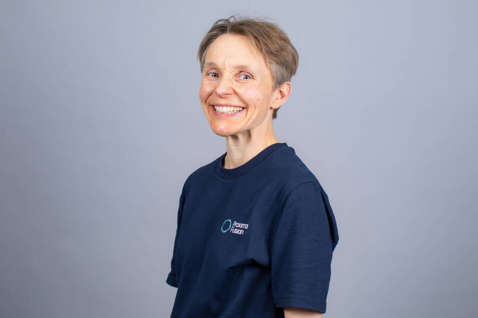 A headshot photo of Jenny Cane - the background is grey with Jenny stood in the centre with a big smile
