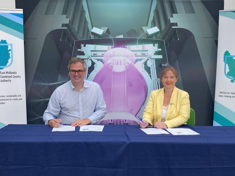 A man holding a pen sat on the left in front of a table with two sheets of paper  in front of him with a woman sat on the right also holding a pen sat in front of the table with two sheets in front of her