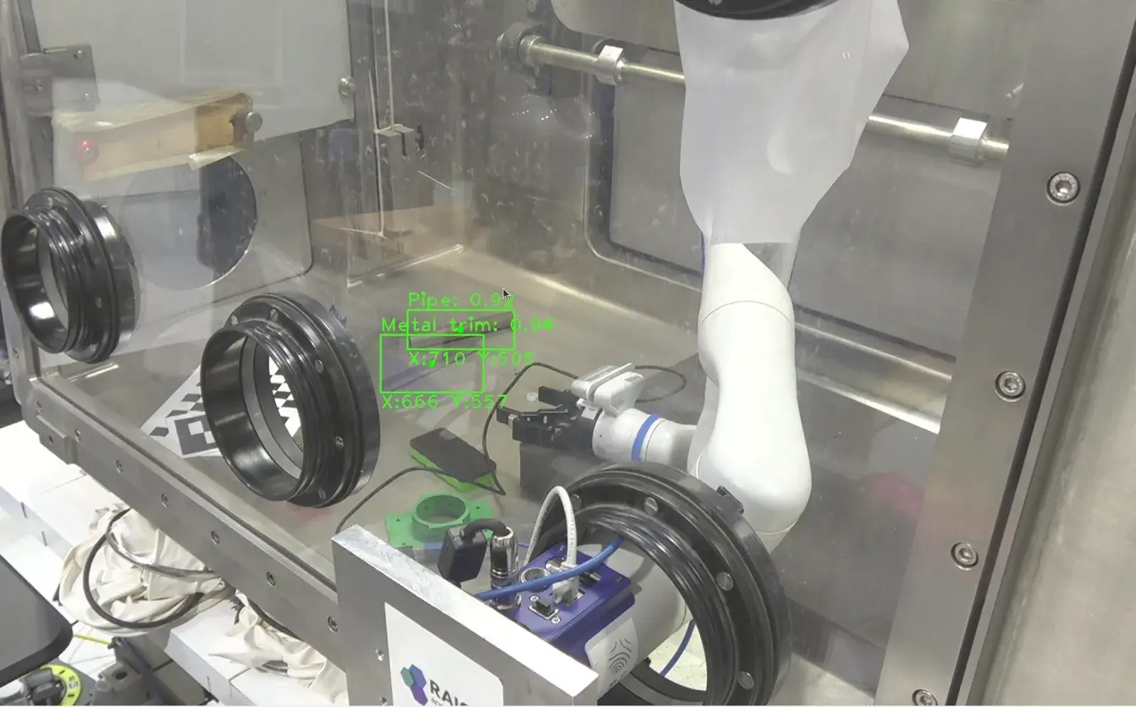 A large metal box with a glass side that has robotic arms in side along with a metal ruler