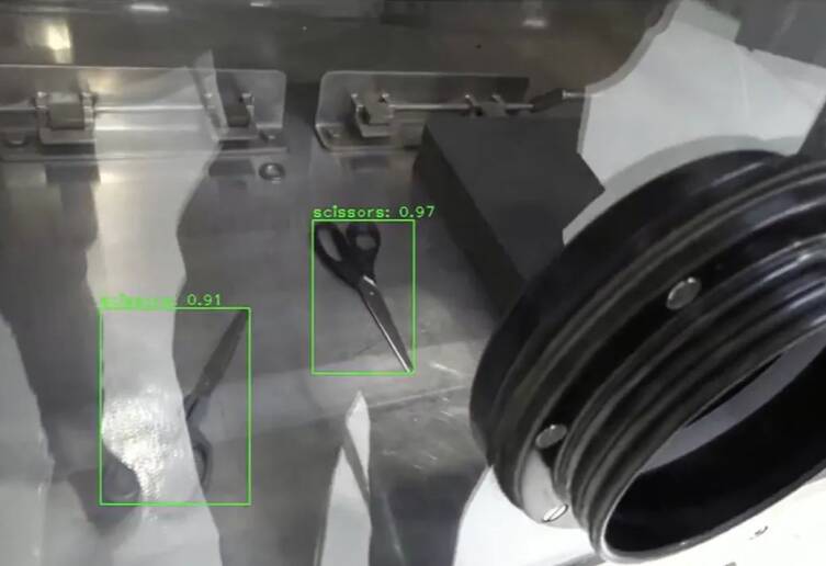 A close up of a large metal box with a glass side that has robotic arms in the box is also scissors