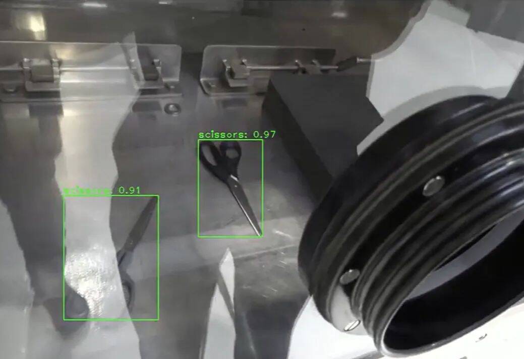 A close up of a large metal box with a glass side that has robotic arms in the box is also scissors