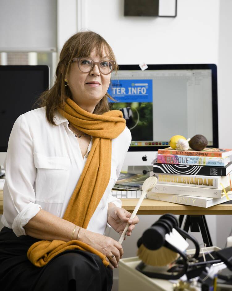 Foto van ontwerpster Brigitte Beenen die tevens een fervente kok is. Op de foto zit Brigitte met een pollepel in haar hand voor haar computer met naast haar een stapel kookboeken.
