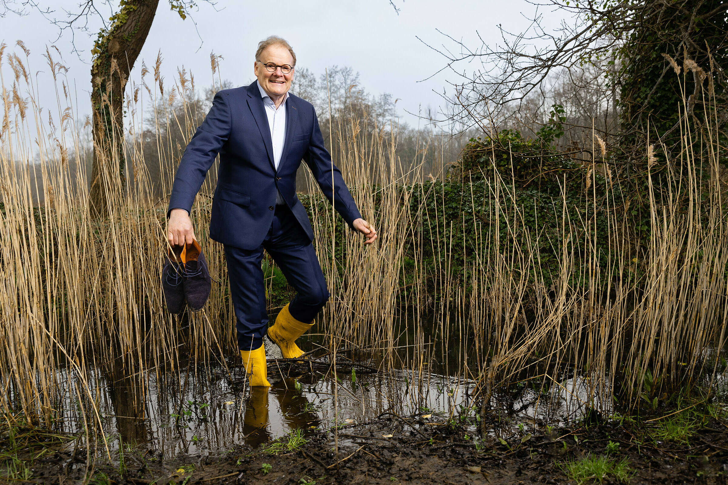 Foto van Joost Buntsma die in een blauw pak en met gele laarzen aan uit een vennetje in een waternatuurgebied stapt. Hij draagt zijn nette schoenen in zijn hand.