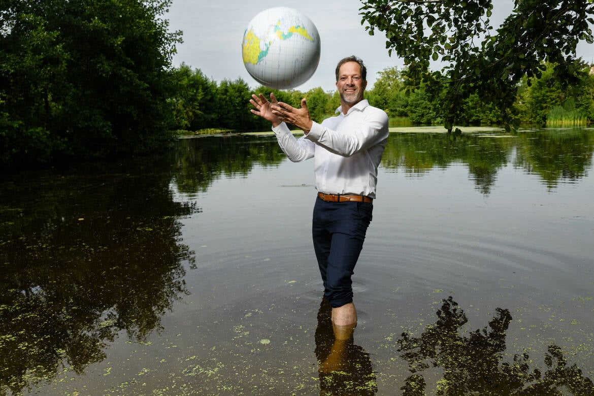 Foto van Mark van der Werf, de nieuwe directeur van STOWA, met een opblaasbare wereldbol in zijn handen terwijl hij met blauwe broek en wit overhemd in een meertje staat.