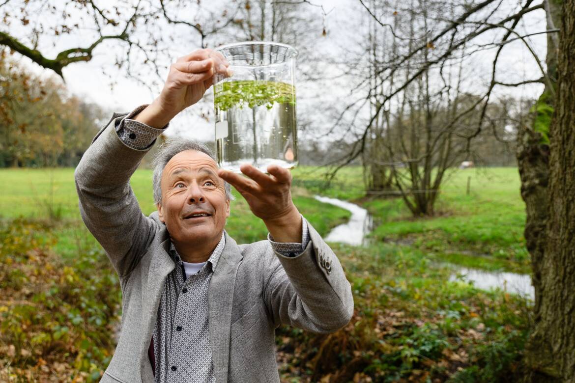 Foto van Harm Gerrits in een beeklandschap terwijl hij die een glazen maatbeken boven zijn hoofd houdt met daarin water en een waterplant.