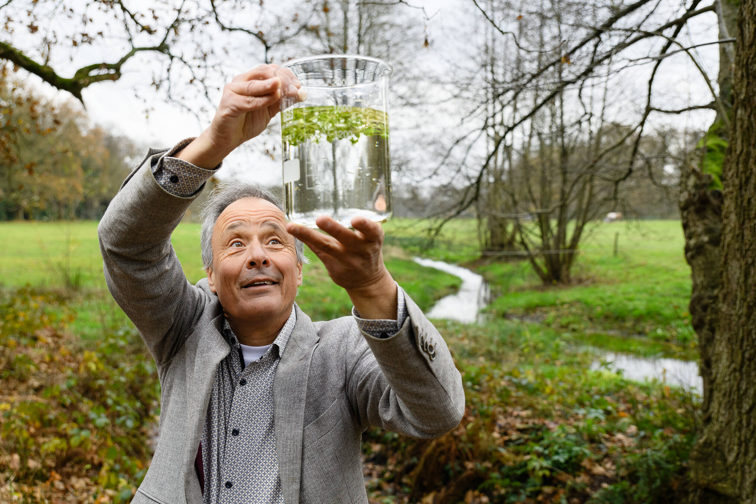 Foto van Harm Gerrits in een beeklandschap terwijl hij die een glazen maatbeken boven zijn hoofd houdt met daarin water en een waterplant.