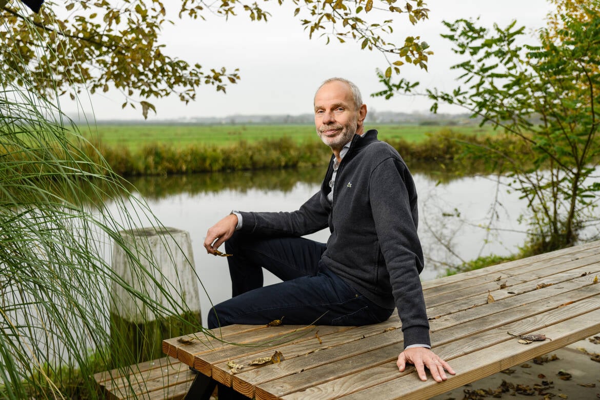 Foto van Jan Willem Erisman zittend op een houten bank voor een sloot.