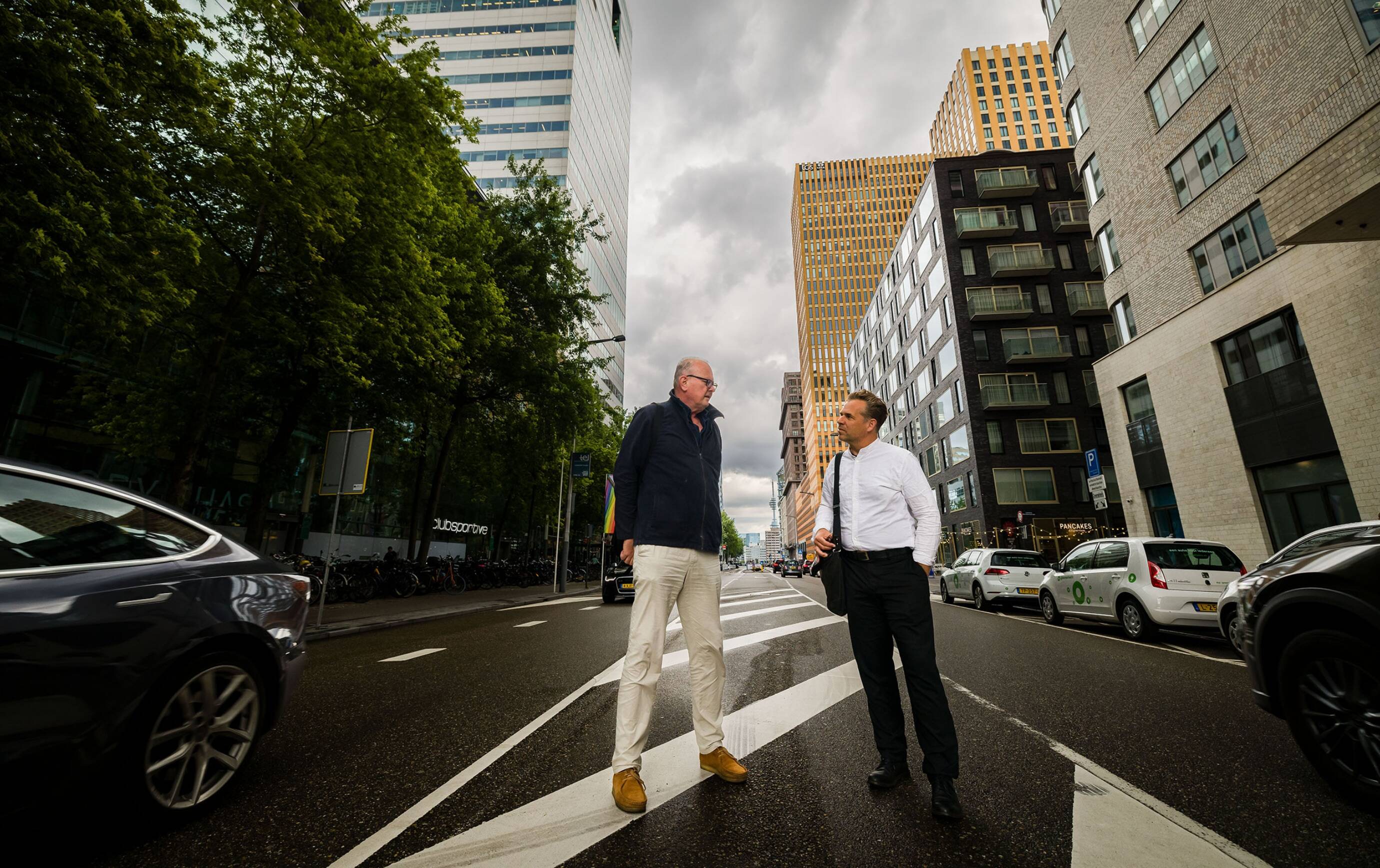 Portret van Ton Schaap en Paco Bunnik die elkaar geïnteresseerd aankijken, op straat tussen auto’s en hoge gebouwen