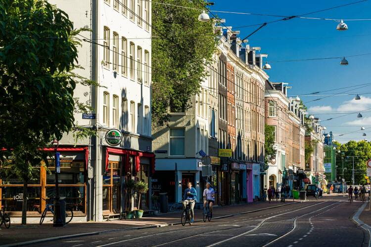 Een zonnige Ferdinand Bolstraat ter hoogte van Brasserie Lolita, met historische gevels, kleurige plinten, fietsers en voetgangers.