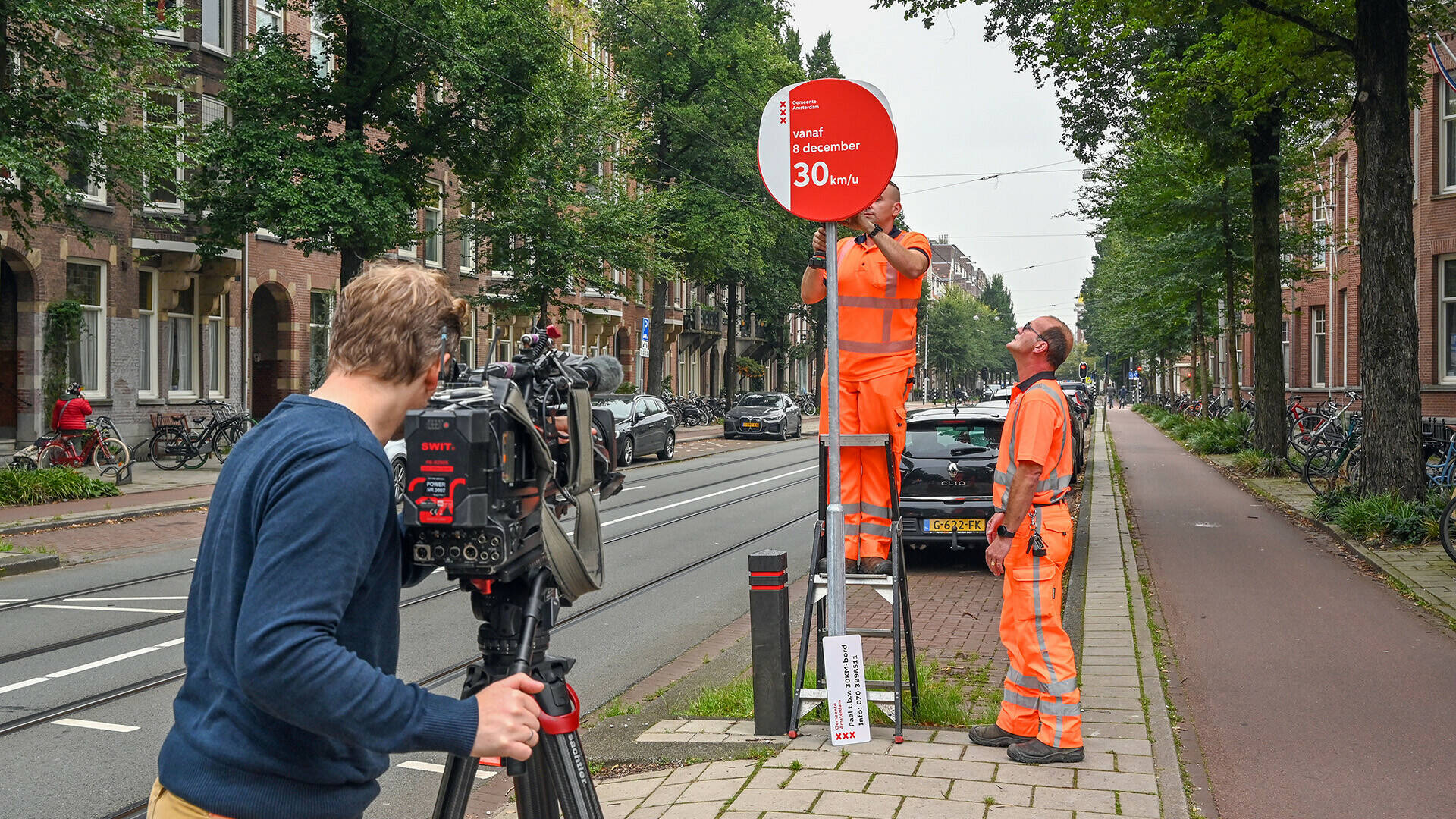 Medewerkers van de gemeente plaatsen een bord met 30 km per uur. Op de voorgrond een cameraman