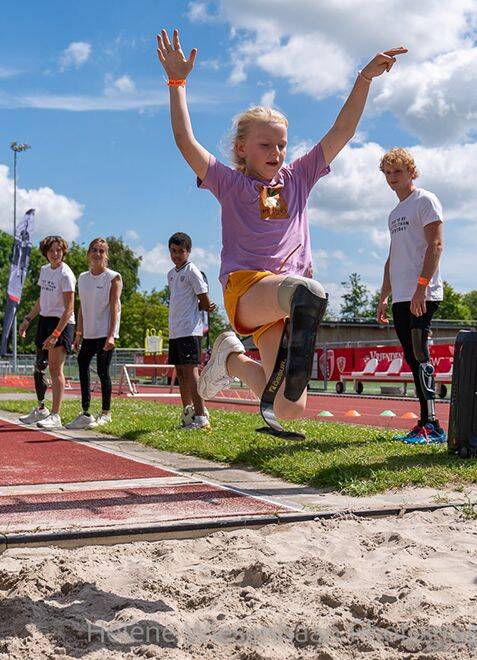 Een jong kind met een prothese maakt een sprong tijdens een atletiektraining op een zonnige dag.