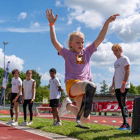 Een jong kind met een prothese maakt een sprong tijdens een atletiektraining op een zonnige dag.