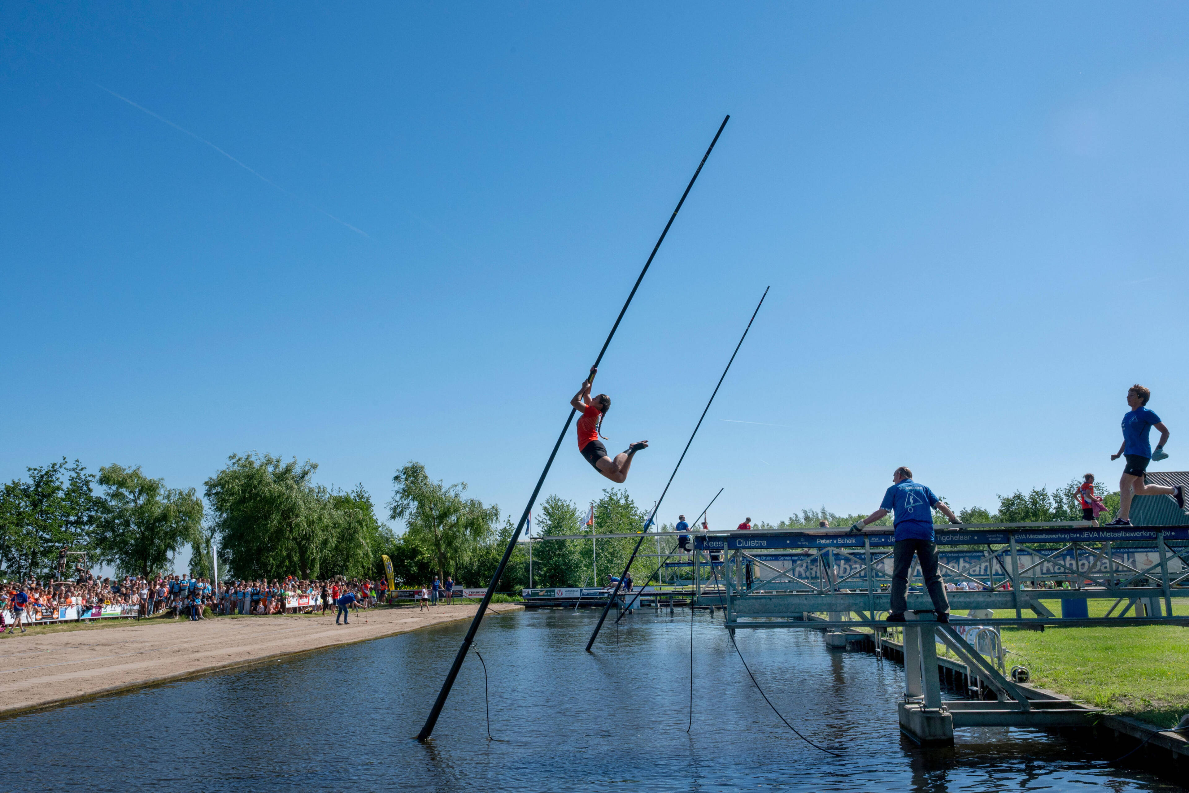 Een vrouw in het rood zweeft met een lange paal boven het water tijdens het fierljeppen; publiek kijkt toe vanaf de kant