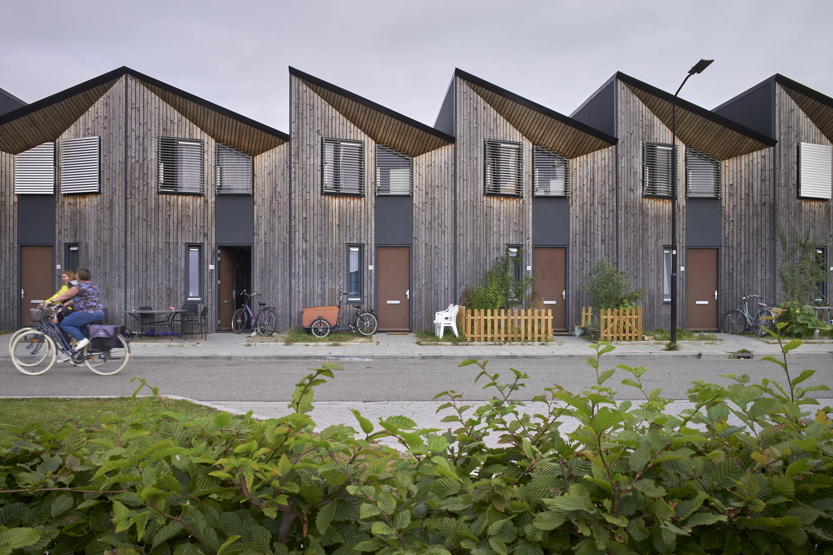 Een vrouw fietst met een kind achterop langs een rij nieuwe woningen met houten gevels en puntdaken