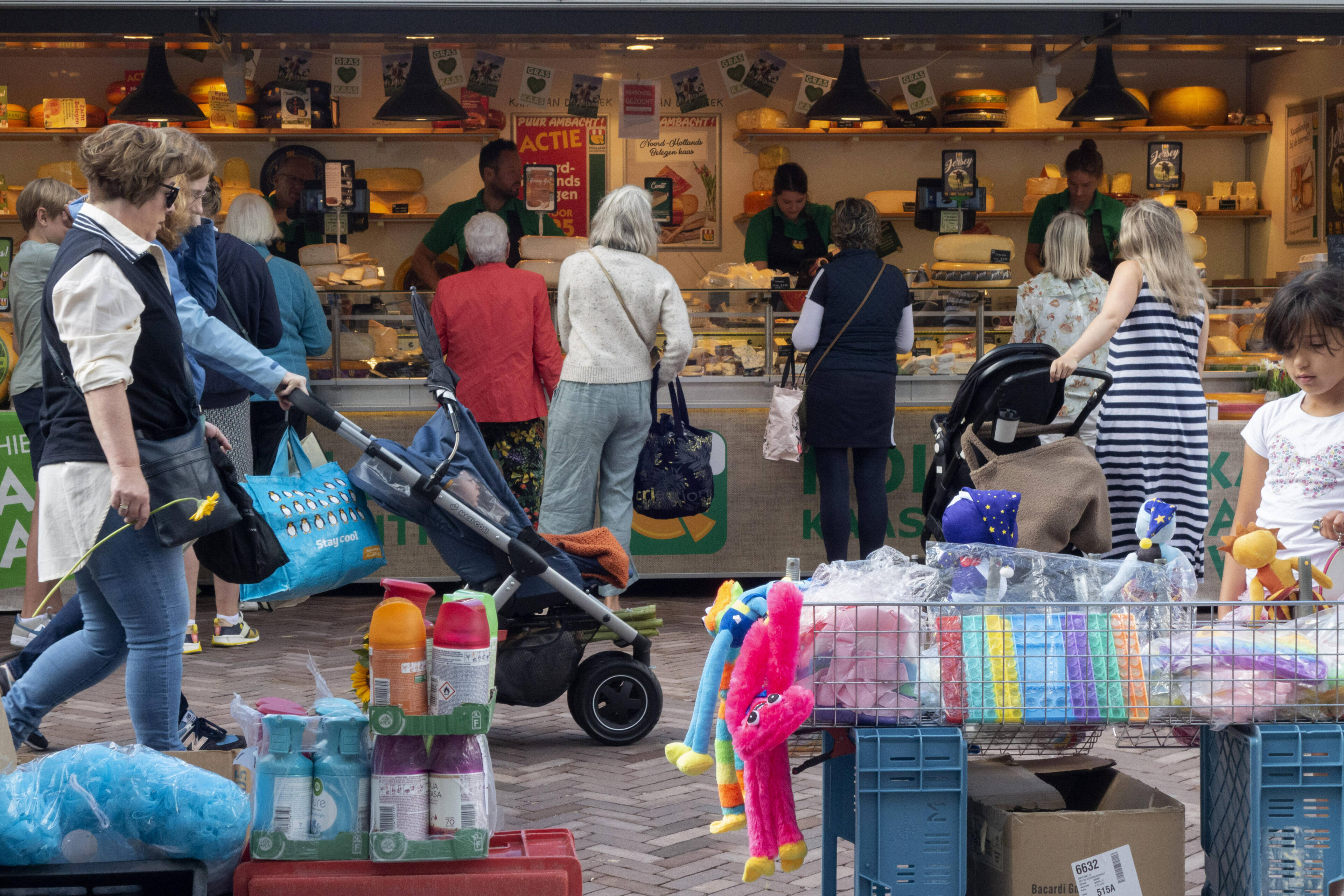 Twee oudere vrouwen praten met elkaar voor een winkel; een van hen zit op een rollator, de winkelruit hangt vol met posters over een uitverkoop.