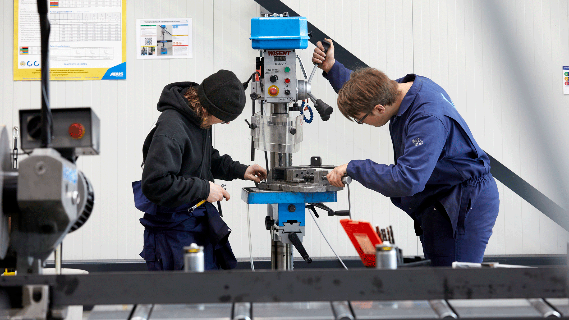 Twee personen in werkkleding werken aan een machine in een techniekopleidingsruimte.
