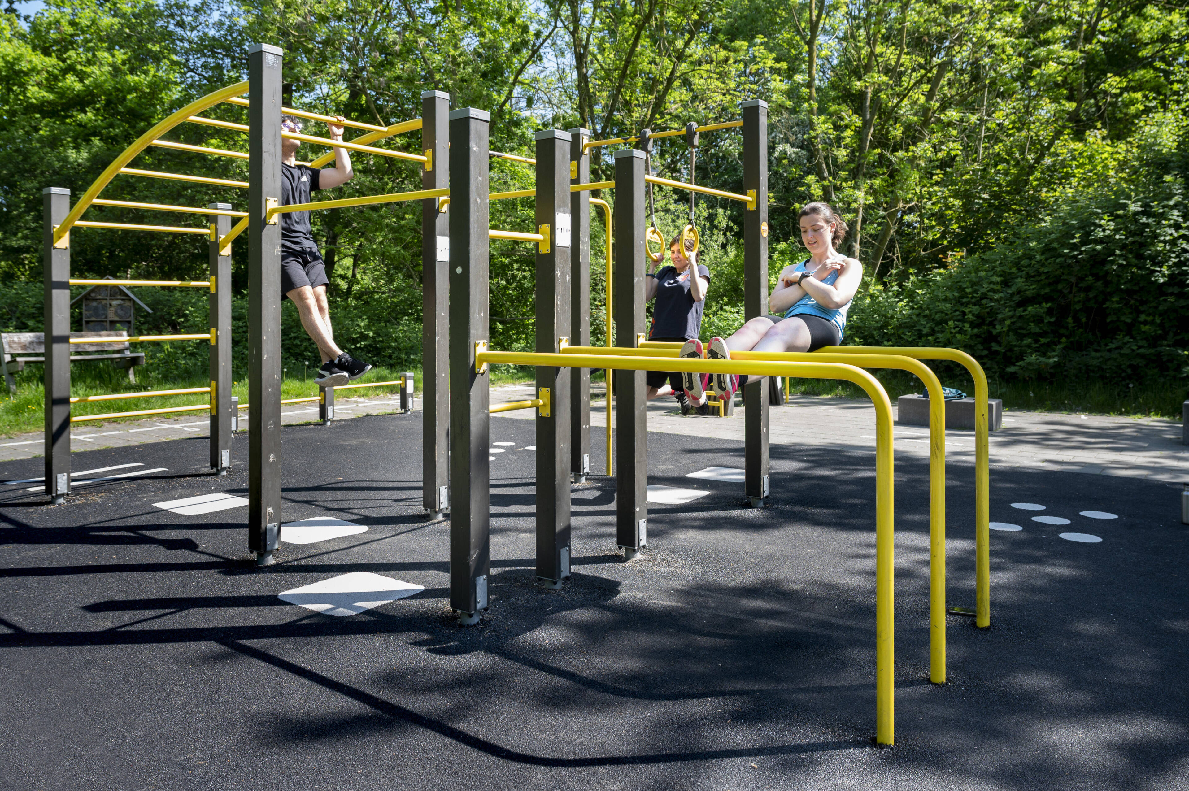 Drie personen sporten op toestellen in een buitenfitnesspark: een man doet pull-ups, twee vrouwen gebruiken de ringen en de lage brug.