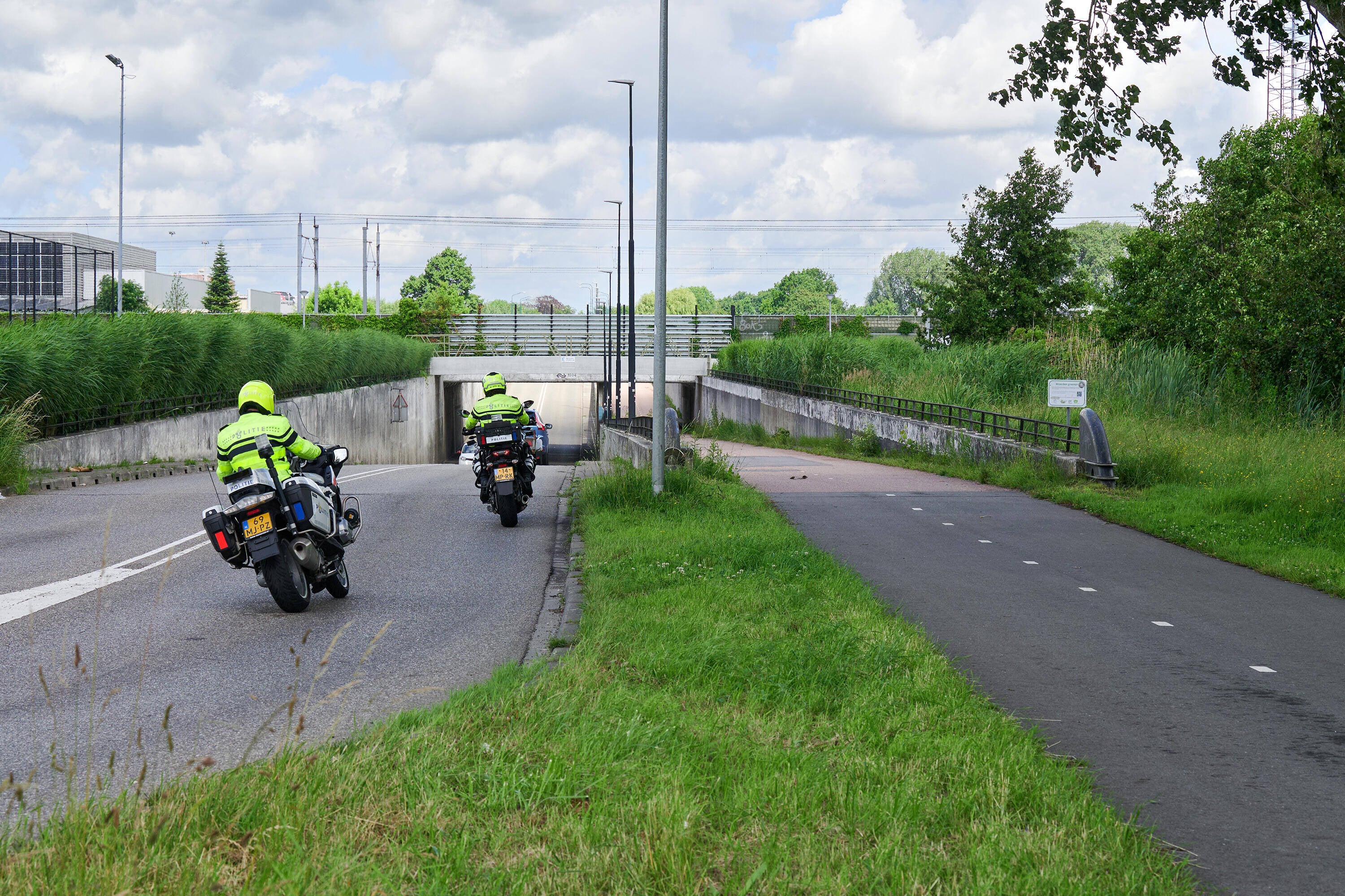Twee politiemotoren met agenten rijden over een weg met fietspad richting een onderdoorgang.