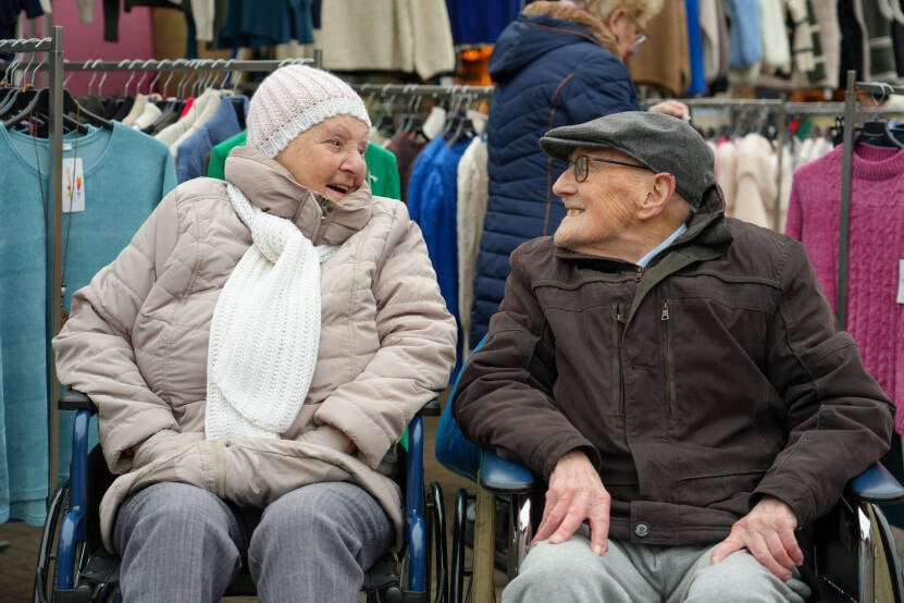 Foto van een oudere vrouw en man zitten naast elkaar in een rolstoel die gezellig kletsen.