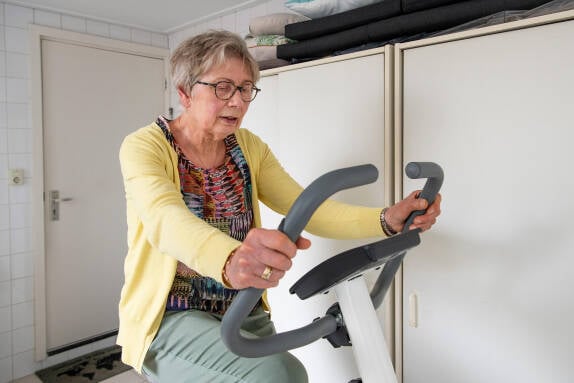 Foto van een oudere mevrouw die in een kamer op een hometrainer zit