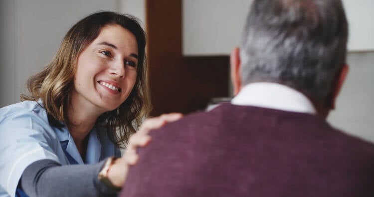Foto van een jonge vrouwelijke zorgmedewerker die haar hand geruststellend op de schouder van een oudere meneer legt.