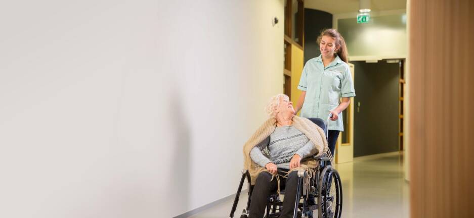 Foto van een oudere mevrouw in een rolstoel. Ze kijkt lachend naar de zorgmedewerker die haar duwt.