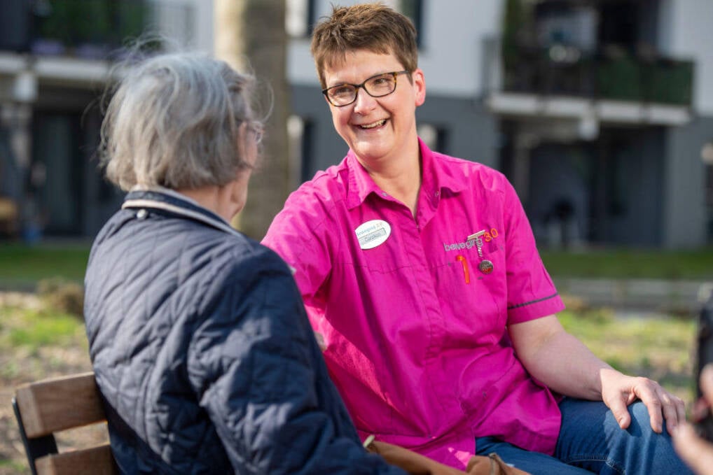 Foto van een zorgmedewerker die met een zorgpati&euml;nt buiten op een bankje zit. Ze lachen naar elkaar.