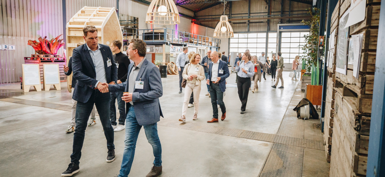 Een groep zakelijk geklede mensen lopen door een agrarische bedrijfshal die omgebouwd is tot evenementen locatie. Twee mannen die vooraan in de groep lopen schudden elkaar de hand. Het lijkt of ze elkaar zo wandelend ontmoeten.