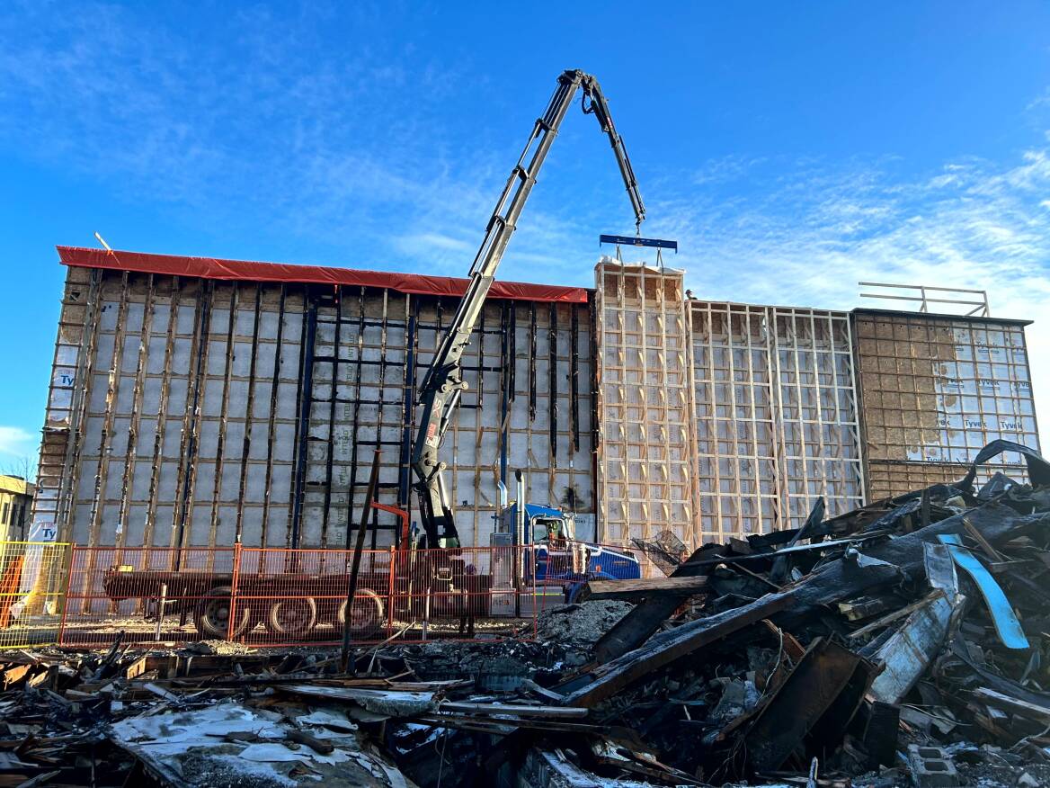 A crane carries a prefab panel in front of a damaged building