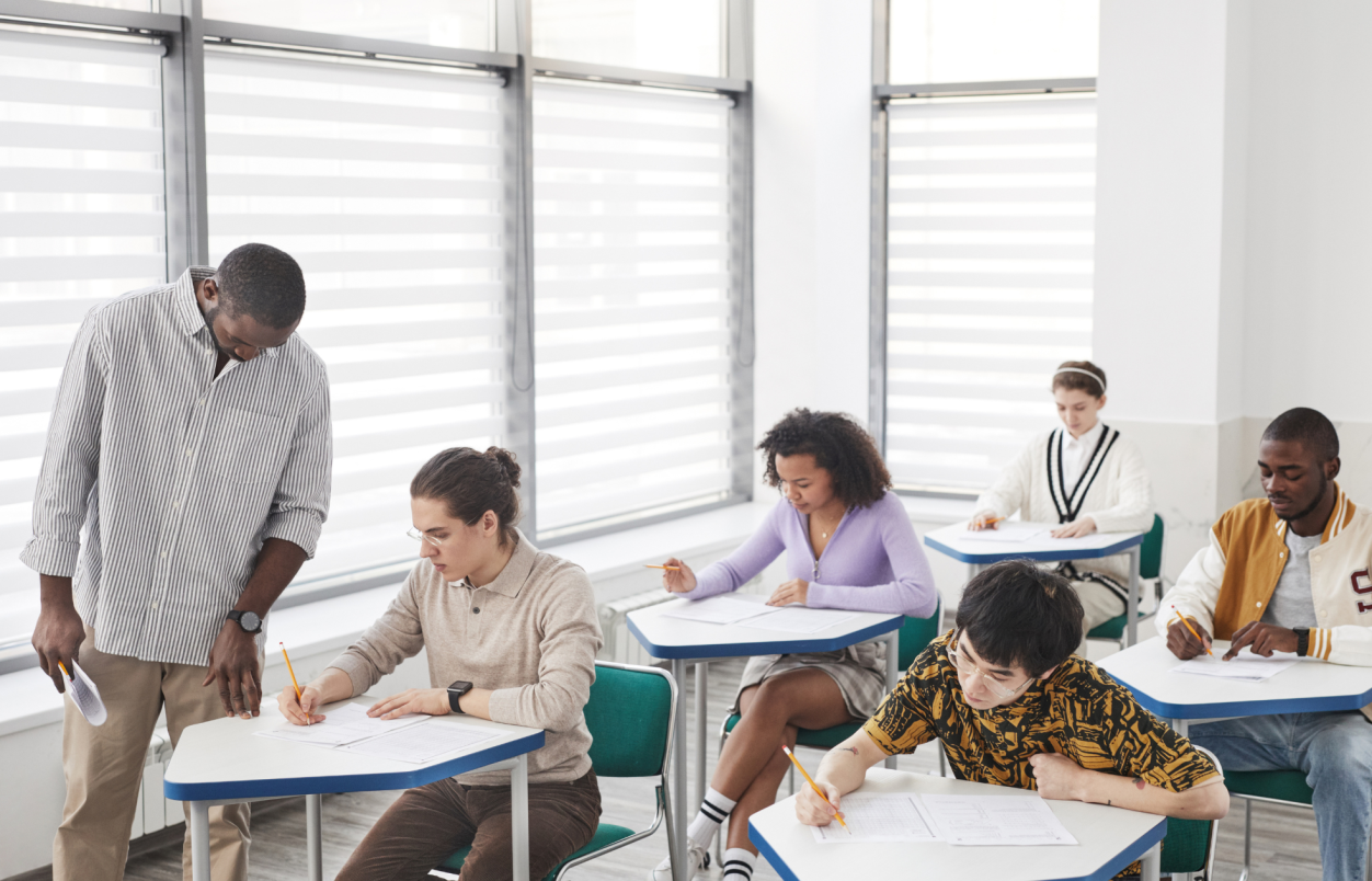 docent kijkt met student die in een klas zit waar een toets wordt afgenomen. De studenten zitten aan afzonderlijke tafels