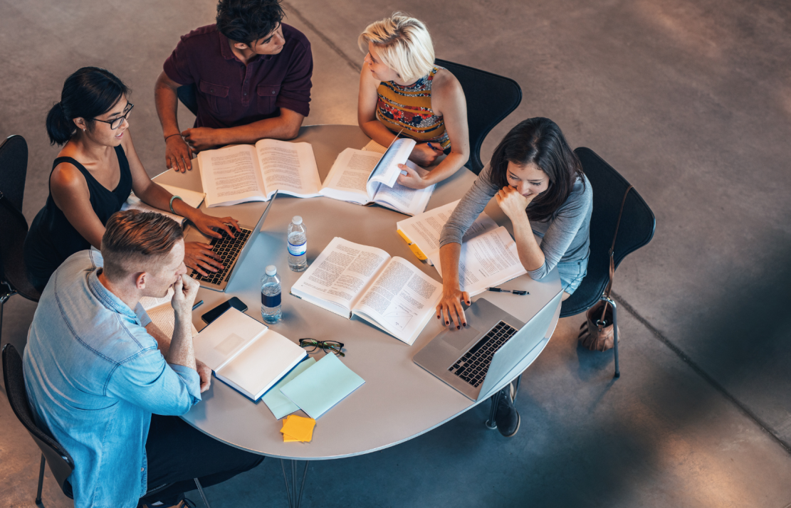 personen aan een ronde tafel in overleg, van bovenaf bezien. op tafel liggen boeken.