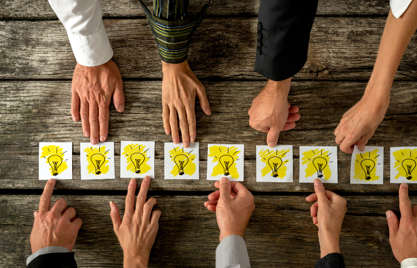 Verschillende handen van diverse personen wijzen naar kaarten met daarop illustraties van gele gloeilampen, die op een houten tafel liggen. De gloeilampen symboliseren idee&euml;n of inspiratie.