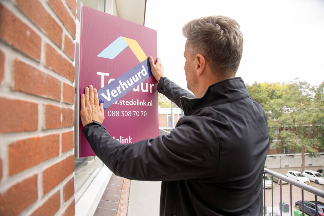 We zien een een medewerker een 'verhuurd' sticker plakken op een makelaarsbord.