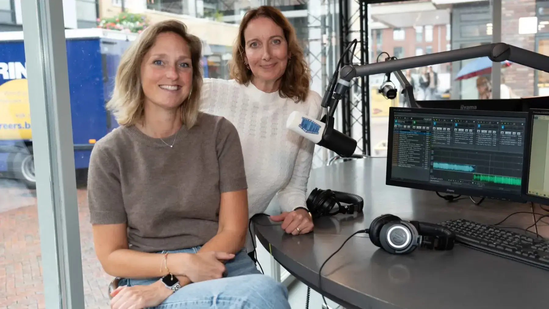 Twee communicatieadviseurs zitten in een radiostudio met microfoon, koptelefoons en een scherm met audioprogramma, terwijl ze vertellen over toegankelijke communicatie.