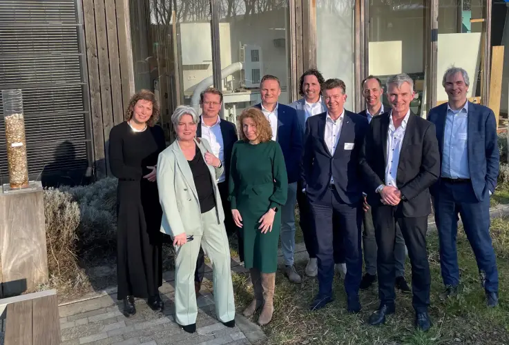Groep samenwerkingspartners staat buiten bij gebouw en markeert eerste jaar samenwerking in renovatieproject.