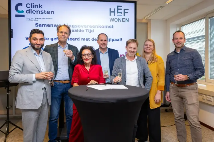 Groep medewerkers en partners staat rond tafel met ondertekeningsdocument en heft glas tijdens offici&euml;le samenwerkingsovereenkomst.