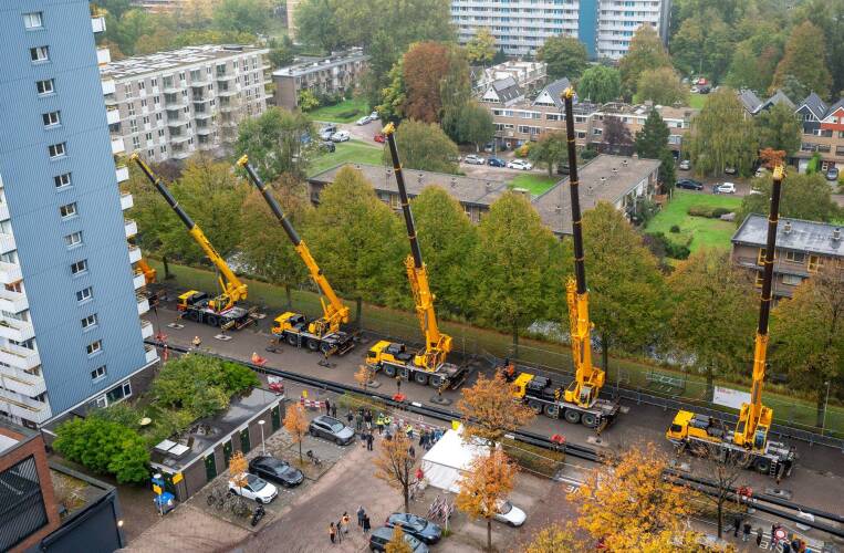 Luchtfoto van werkzaamheden met meerdere grote gele kranen die lange leidingen plaatsen langs woongebouwen voor aanleg van warmtenet.
