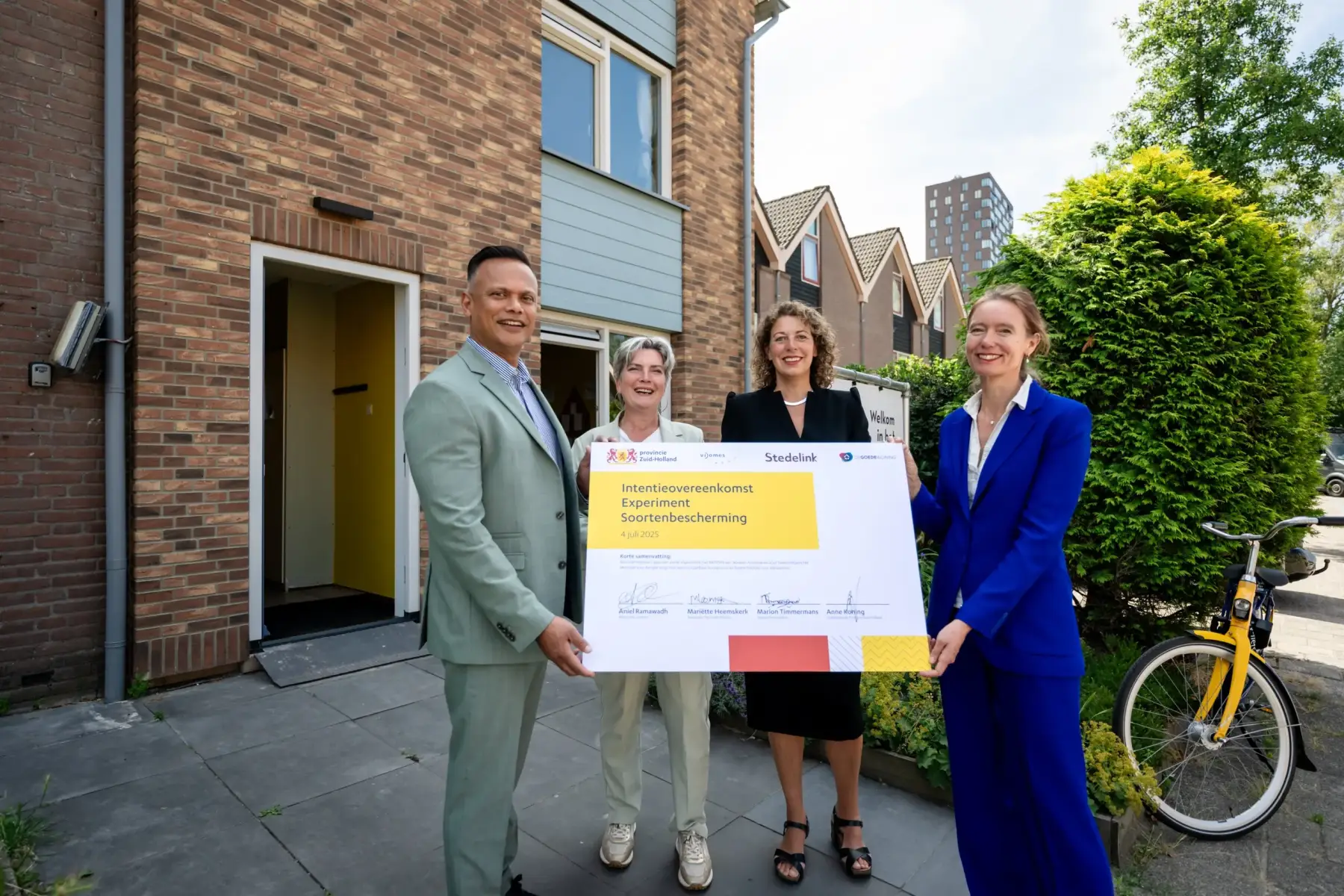 Vier vertegenwoordigers staan voor een woongebouw en tonen een ondertekende overeenkomst voor soortenbescherming.