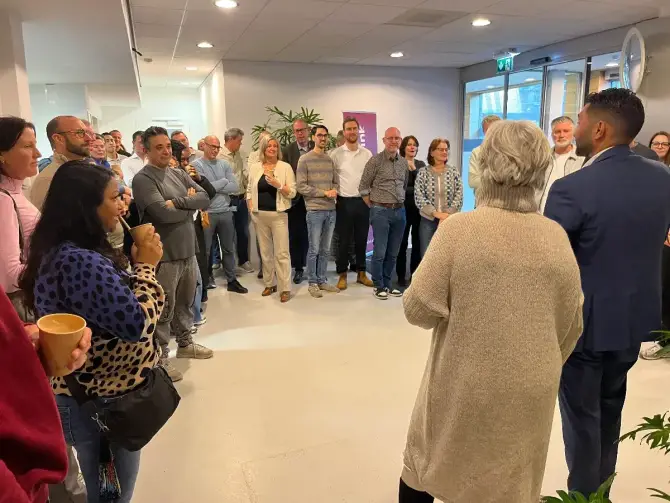 Collega&rsquo;s staan samen in kantoorruimte en luisteren naar presentatie tijdens bijeenkomst over organisatieverandering.