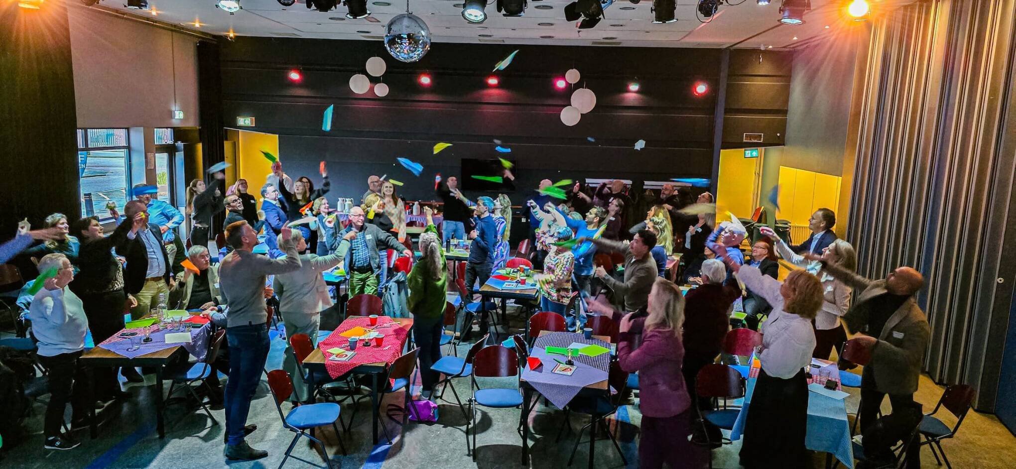 Foto van de bijeenkomst, bezoekers staan in de ruimte en gooien zelf gevouwen papieren vliegtuigjes in de lucht.