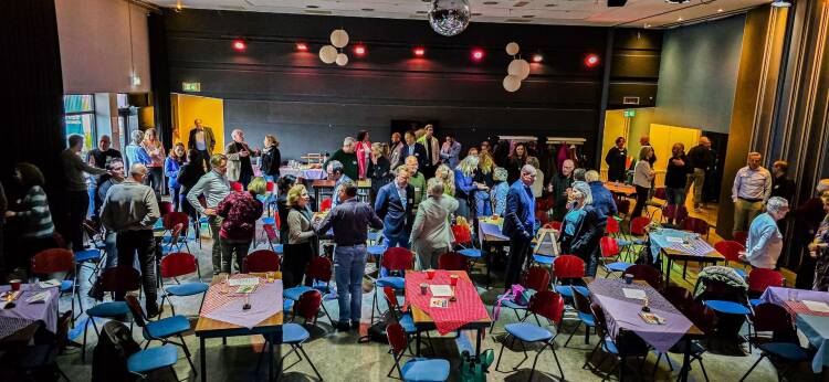 Foto van de bijeenkomst, bezoekers staan in groepjes bij elkaar te overleggen.