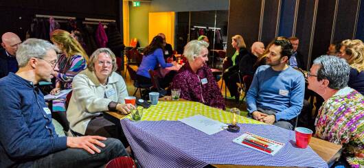 Foto van de bijeenkomst, bezoekers overleggen met elkaar aan tafels.