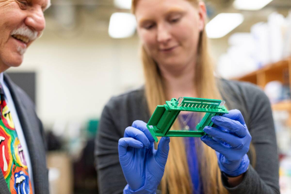 A lab staff member with gloves on is holding a research sample and showing it to Dr. Herb Paaren.