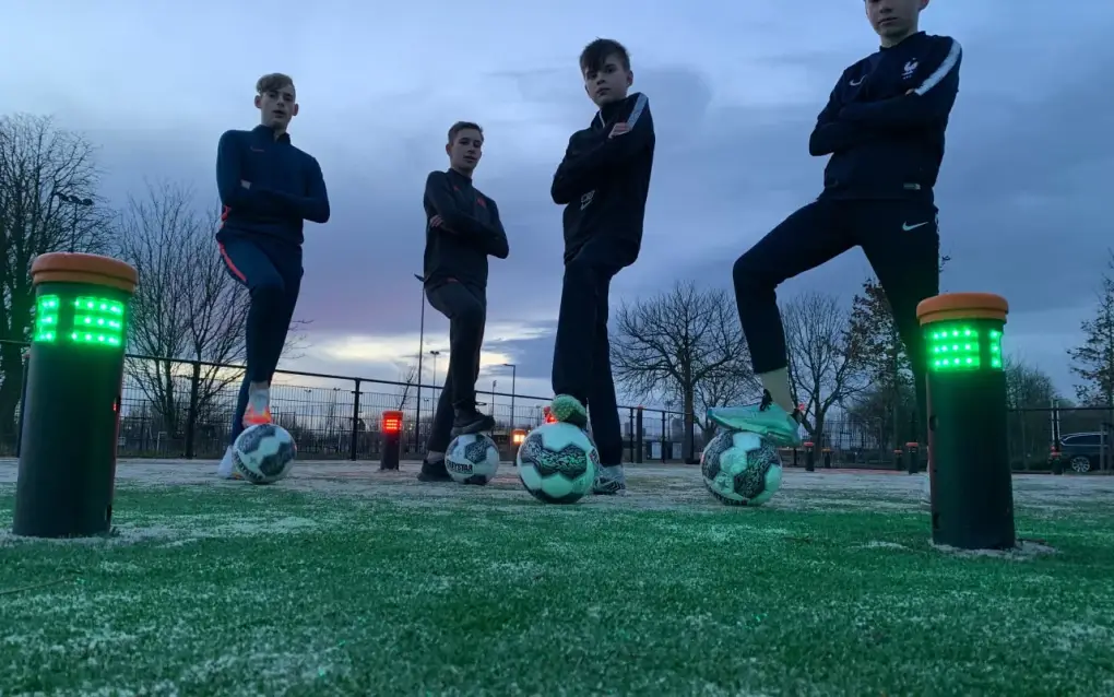 Vier mannen staan naast verlichte interactieve voetbalpalen op een kunstgrasveld.